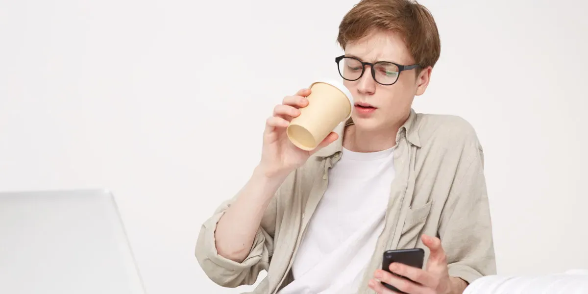 Teenage boy drinking coffee looking at phone with books around