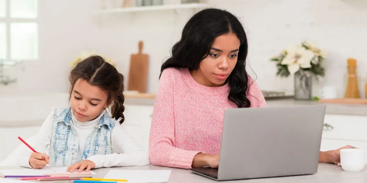 Mother working on laptop with daughter drawing beside her