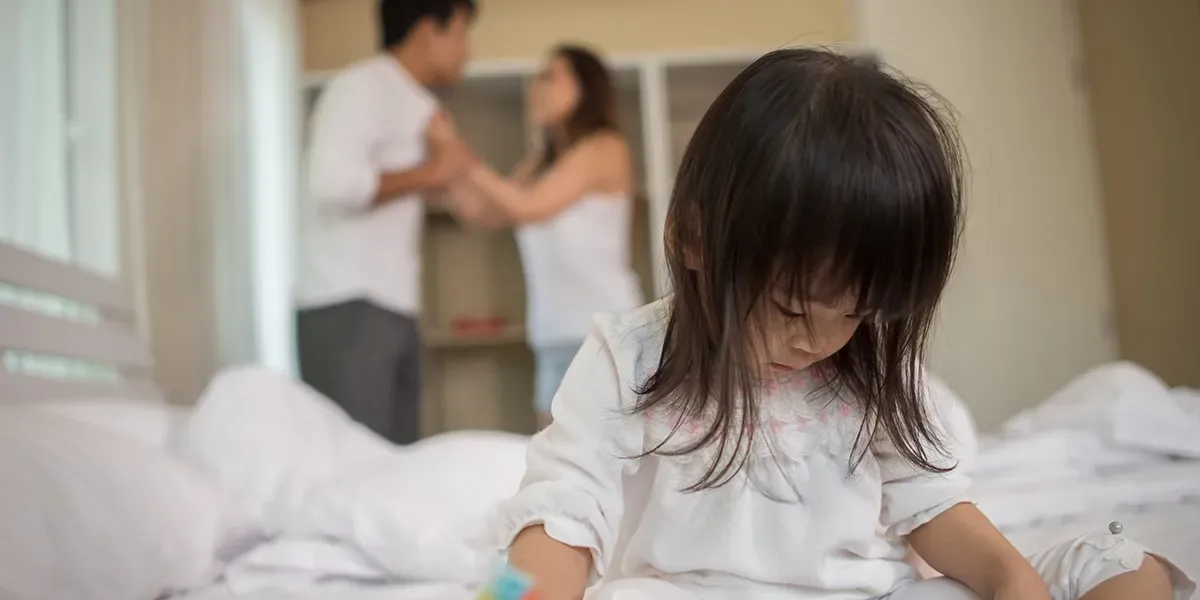 Young girl sitting on bed with parents fighting in the background